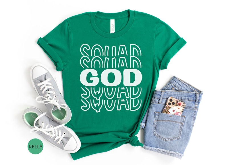 Pu&ograve; includere: Una maglietta verde con il testo "GOD SQUAD" in lettere bianche con un design retr&ograve;. La maglietta &egrave; stesa su uno sfondo bianco con un paio di scarpe da ginnastica grigie e pantaloncini di jeans blu.