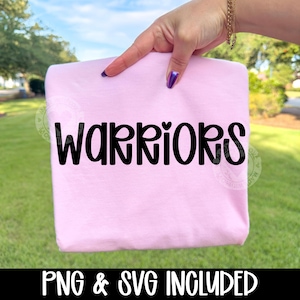 May include: A light pink t-shirt with the word "WARRIORS" in bold black letters. The text "PNG & SVG INCLUDED" is printed in black at the bottom of the shirt. The shirt is held up by a hand.