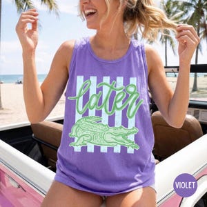 May include: A violet tank top with a green alligator graphic and the word "later" in a cursive font. The text and alligator are set against a backdrop of vertical light blue stripes. The tank top is sleeveless.
