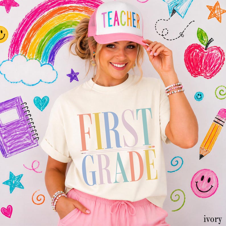 May include: A cream-colored t-shirt with the text FIRST GRADE in multicolored letters and a matching pink and white trucker hat with the word TEACHER in multicolored letters. The shirt features a pencil graphic underlining the word FIRST.