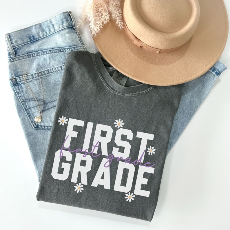 Op de afbeelding: Een donkergrijs T-shirt met de tekst "FIRST GRADE" in witte letters met een paarse "first grade" eronder. Het shirt is versierd met witte madeliefjes.
