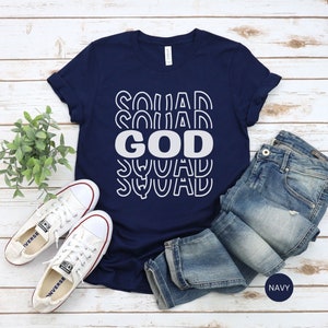 Pu&ograve; includere: Una maglietta blu navy con la scritta "GOD SQUAD" stampata in bianco. Il testo &egrave; ripetuto in un design retr&ograve; impilato. La maglietta &egrave; stesa su una superficie di legno con un paio di scarpe da ginnastica bianche e un paio di jeans blu.
