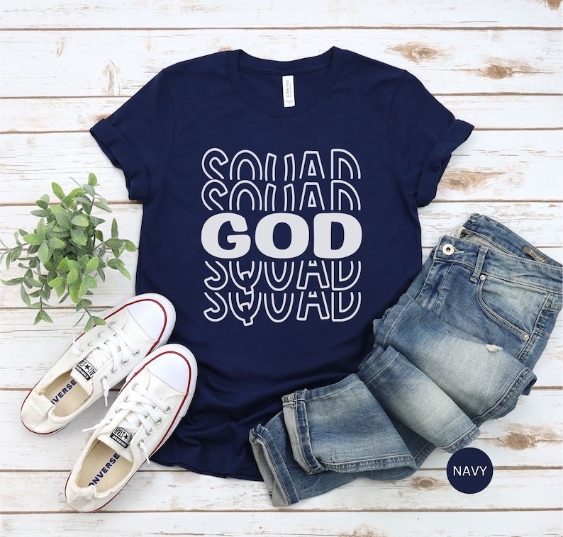Pu&ograve; includere: Una maglietta blu navy con la scritta "GOD SQUAD" stampata in bianco. Il testo &egrave; ripetuto in un design retr&ograve; impilato. La maglietta &egrave; stesa su una superficie di legno con un paio di scarpe da ginnastica bianche e un paio di jeans blu.