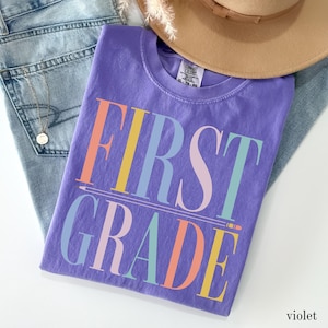 May include: A violet short-sleeve t-shirt featuring the text FIRST GRADE in large, multicolored block letters. The design includes a horizontal line resembling a pencil underline. The shirt has a crew neckline and a white interior label.