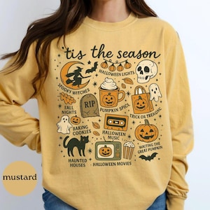 May include: Mustard-colored long-sleeve shirt with a Halloween-themed graphic. The design includes a witch on a crescent moon, pumpkins, a skull, and the text "'tis the season". Other elements include a black cat, ghost, and the words "Spooky Witches" and "Haunted Houses".