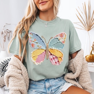 May include: A light green t-shirt featuring a colorful butterfly applique. The butterfly design incorporates various fabric patterns, including floral, gingham, and solid colors. The word "bay" is visible at the bottom left of the image.