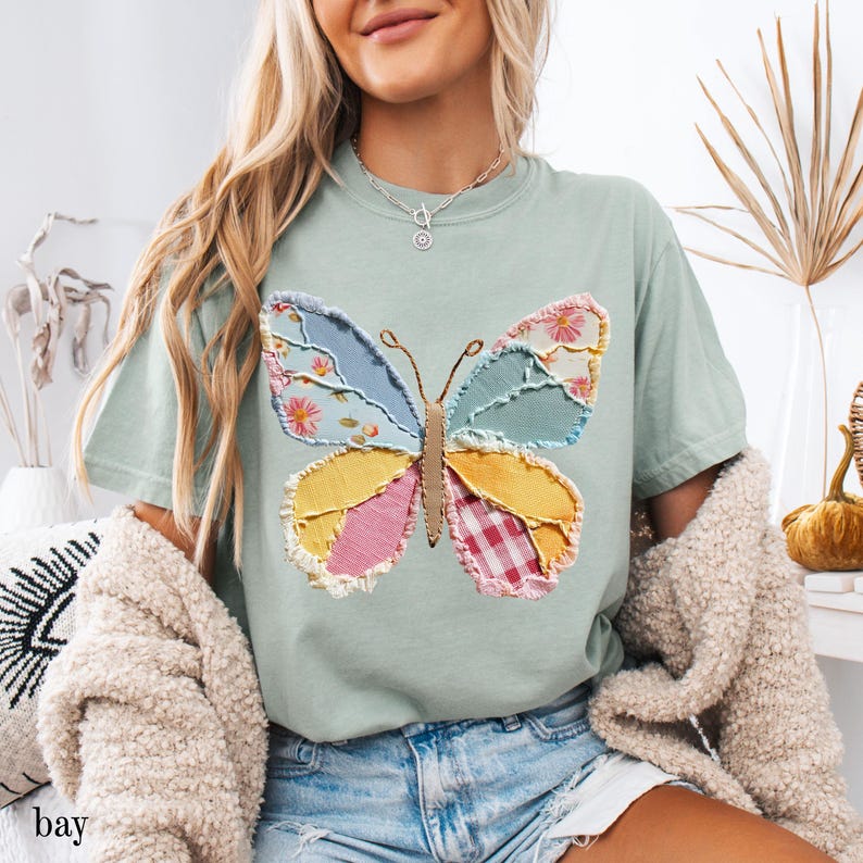 May include: A light green t-shirt featuring a colorful butterfly applique. The butterfly design incorporates various fabric patterns, including floral, gingham, and solid colors. The word "bay" is visible at the bottom left of the image.