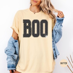 Puede incluir: Una camiseta de manga corta de color amarillo p&aacute;lido con la palabra BOO impresa en grandes letras negras con un patr&oacute;n de lunares blancos. La camiseta se lleva con una chaqueta vaquera azul sobre los hombros. El texto BUTTER aparece en un c&iacute;rculo.