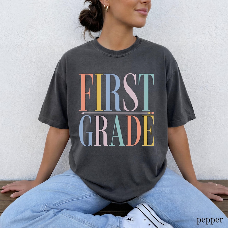 May include: A charcoal gray short-sleeve t-shirt featuring the text FIRST GRADE in a multi-colored serif font. The word FIRST is stacked above the word GRADE, separated by a horizontal line graphic resembling a pencil with a pointed tip.