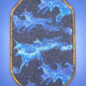 May include: A rectangular wall hanging with a celestial design. The artwork features blue unicorns against a dark blue and black starry background. The frame is a light brown color with a decorative pattern, and a brass-colored metal loop is at the top for hanging.
