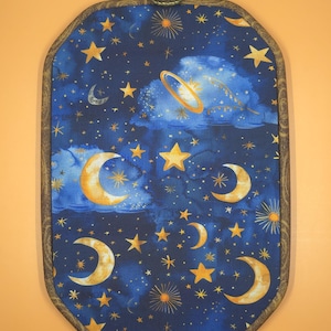 May include: An oval-shaped wall hanging featuring a celestial design. The fabric displays a dark blue background with gold moons, stars, and a ringed planet. The wooden frame has a decorative edge and a metal handle at the top.