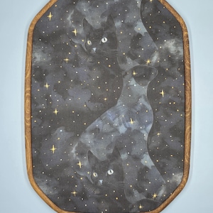 May include: An oval-shaped wall hanging featuring a celestial design with black cats and a starry night sky. The artwork is framed in a wooden border with a brass-colored clasp at the top. The cats have white eyes and are set against a dark background.