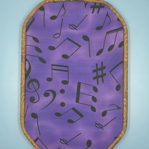 May include: A decorative, framed piece featuring a purple fabric background adorned with black musical notes and symbols. The frame is a warm, patterned wood with a brass-colored clasp at the top. The overall shape is a rounded rectangle.