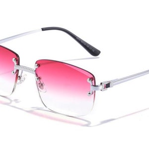 Puede incluir: Gafas de sol con montura plateada y lentes de color rosa degradado. Las gafas tienen un dise&ntilde;o sin montura y una fina montura de metal.