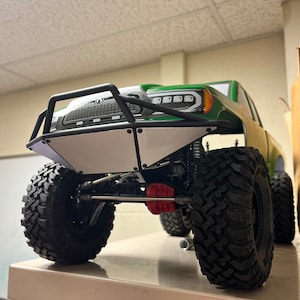 May include: A green and black toy truck with a white front bumper and black wheels. The truck has a black roll cage and a black grille.