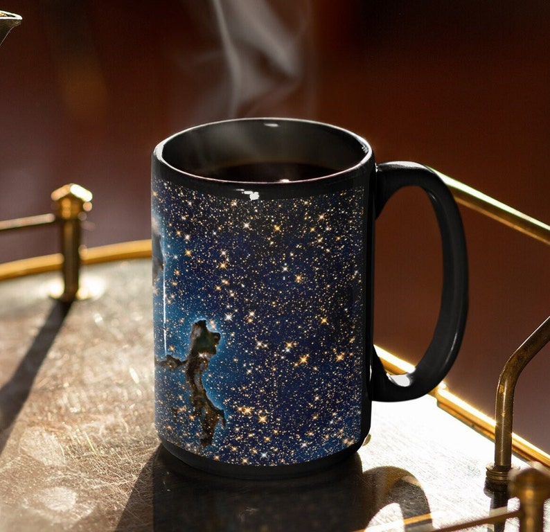 May include: A black coffee mug with a galaxy-themed design. The mug is filled with coffee and steam is rising from the top. The mug is sitting on a gold-colored tray.