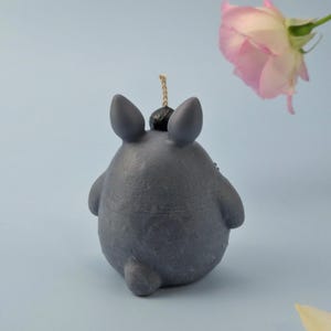 May include: A gray candle shaped like a cartoon character with large ears and a small tail. The candle has a black top and a wick. A pink rose is in the background against a light blue backdrop.