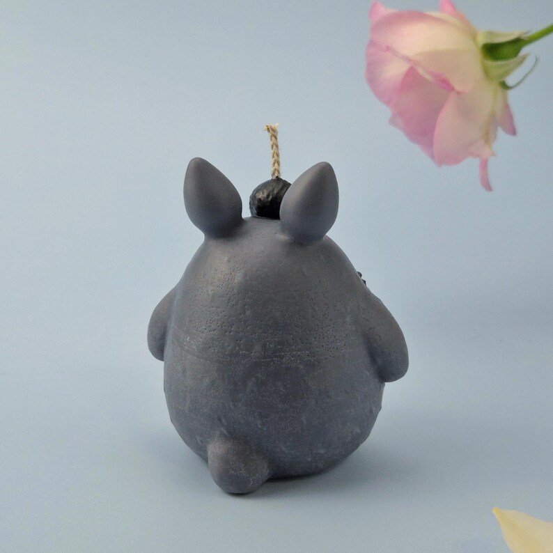 May include: A gray candle shaped like a cartoon character with large ears and a small tail. The candle has a black top and a wick. A pink rose is in the background against a light blue backdrop.