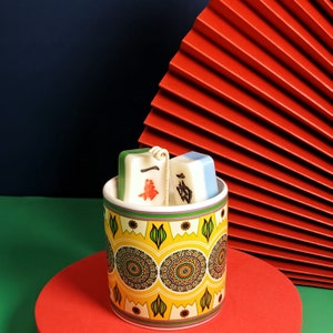 May include: A white ceramic mug with a yellow, green, and brown patterned design. Two white and green ceramic tiles with Chinese characters are sitting inside the mug.