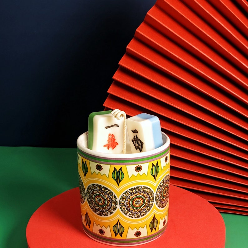May include: A white ceramic mug with a yellow, green, and brown patterned design. Two white and green ceramic tiles with Chinese characters are sitting inside the mug.