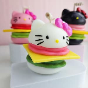 May include: Three Hello Kitty themed candles shaped like hamburgers. Each candle has a different colored Hello Kitty face and a pink bow. The candles are stacked on top of each other, with the bottom candle being green, the middle candle being yellow, and the top candle being pink.