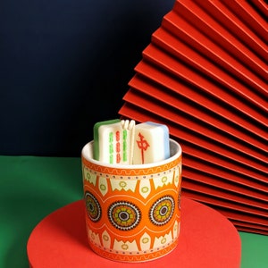 May include: A white ceramic candle holder with a colorful floral pattern. The holder contains two white candles shaped like mahjong tiles. The candles are decorated with green and red markings.