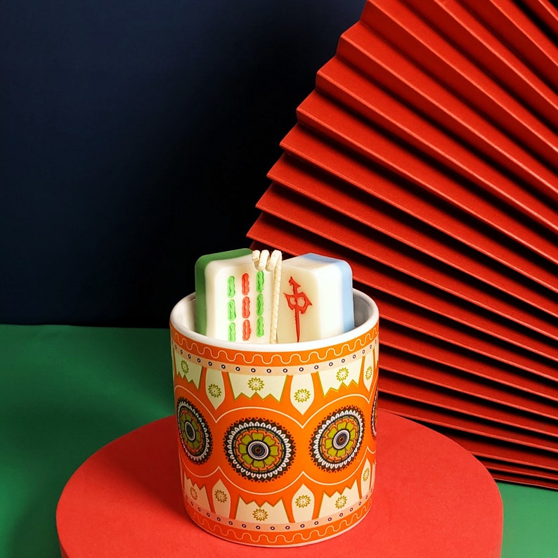May include: A white ceramic candle holder with a colorful floral pattern. The holder contains two white candles shaped like mahjong tiles. The candles are decorated with green and red markings.