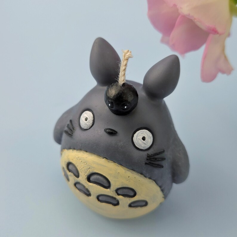 May include: A gray figurine of a cartoon character with large eyes, a yellow belly, and a black top. The figurine has a black sphere on its head with a string attached. A pink flower is in the background.