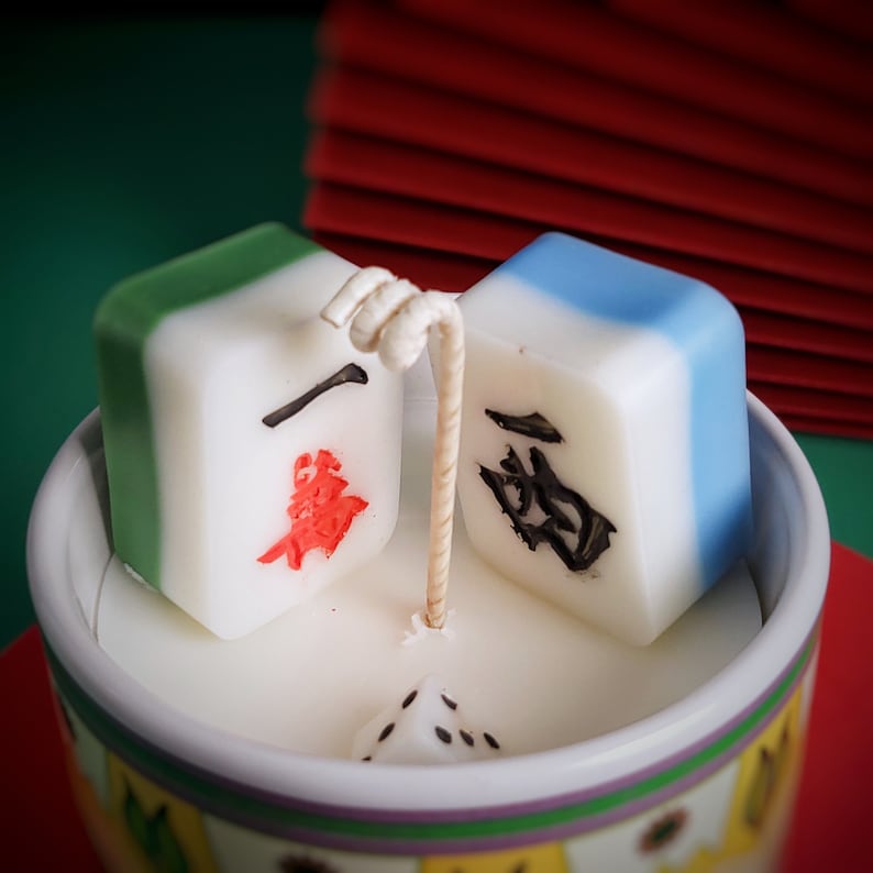 May include: A white ceramic cup with a colorful design around the rim. Inside the cup are three white and colored candles shaped like mahjong tiles. The candles are lit and the wax is melting.