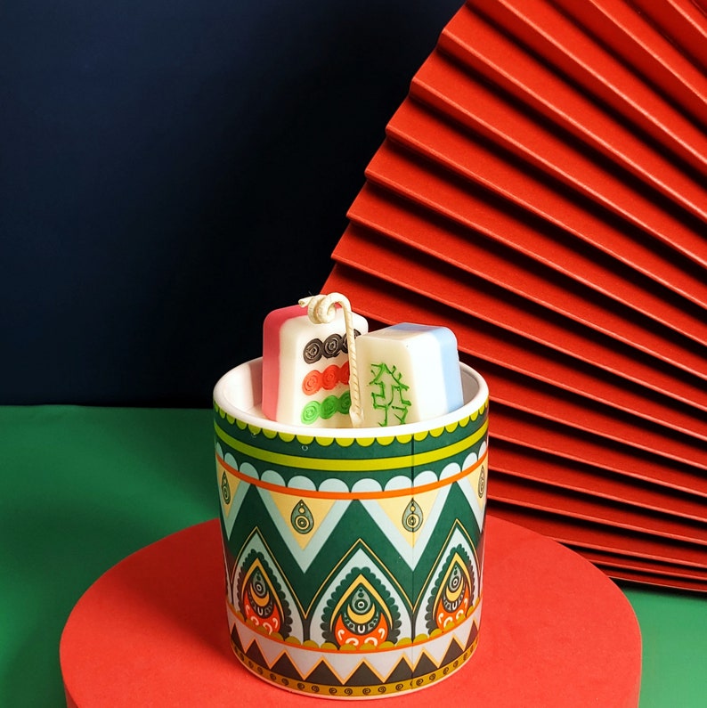 May include: A green and blue ceramic container with a geometric pattern holds three small, white and pink candles shaped like Chinese dominoes. The candles have green and red markings.