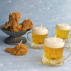 May include: A collection of novelty candles shaped like fried chicken and beer mugs. The chicken candles are a golden brown color and sit in a decorative metal dish. The beer candles have clear glass mugs with a yellow liquid and white foam.