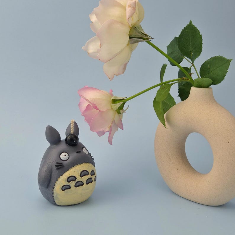 May include: A gray Totoro figurine with a yellow belly and black details sits next to a beige vase with a hole and two pink and white roses. The background is a light blue.