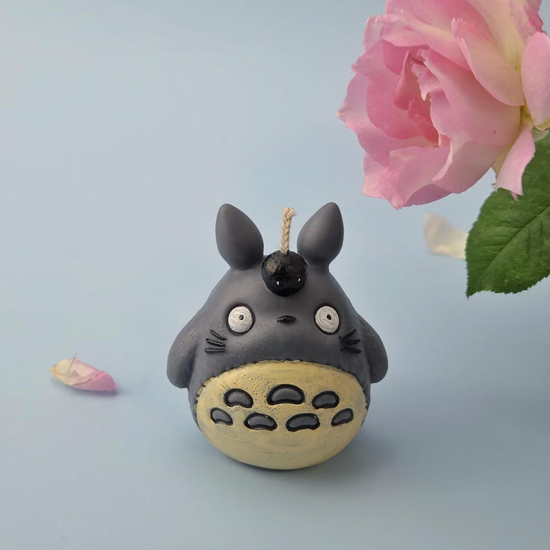 May include: A gray and yellow Totoro-shaped candle with a black top and a wick. The candle has a light blue background with a pink rose and a fallen petal.