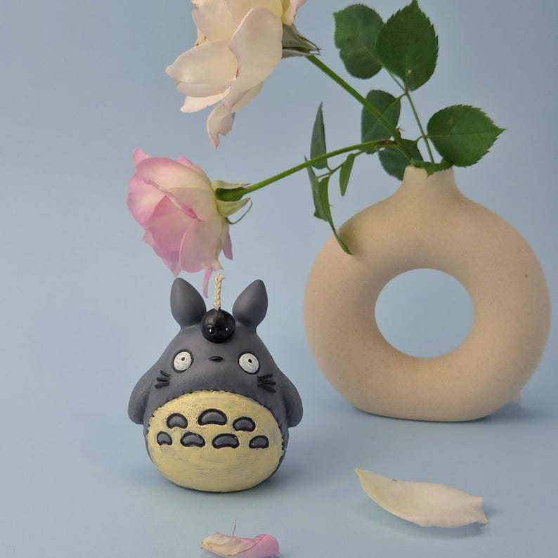 May include: A gray Totoro-shaped candle with a yellow belly and black details. The candle is displayed with pink and white roses and a beige vase against a light blue background.