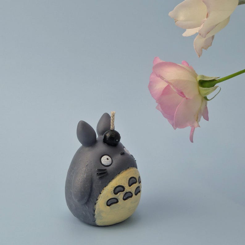 May include: A gray Totoro-shaped candle with a yellow belly and black details. The candle has a wick and a black ball on top. Two roses, one pink and one white, are in the background against a light blue backdrop.