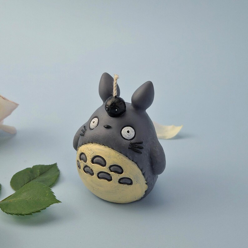 May include: A gray Totoro-shaped candle with a black berry on its head and a beige belly. The candle has large white eyes and small black whiskers. The background is a light blue with green leaves and a white petal.