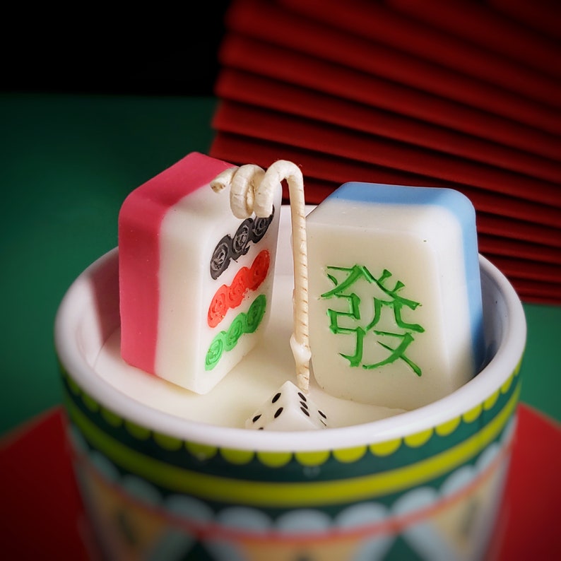 May include: A white ceramic cup with green and red trim, filled with white wax and three colorful candles. The candles are shaped like mahjong tiles, with one red and white tile, one blue and white tile, and one white tile with green Chinese characters. A white dice is also in the cup.