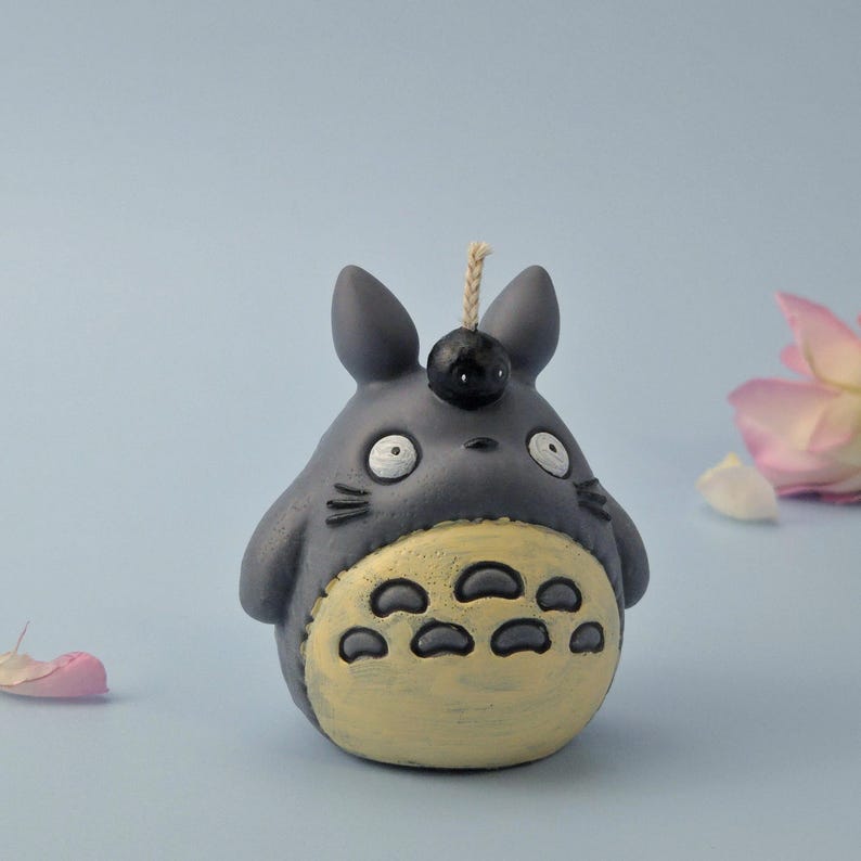 May include: A gray Totoro-shaped candle with a yellow belly and black details. The candle has a wick and a small black ball on top. The background is a light blue with a pink flower and a petal.