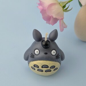 May include: A gray candle shaped like a cartoon character with large eyes, small ears, and a yellow belly. The candle has a black cherry-like detail on its head and a wick. A pink rose and a beige vase are in the background.