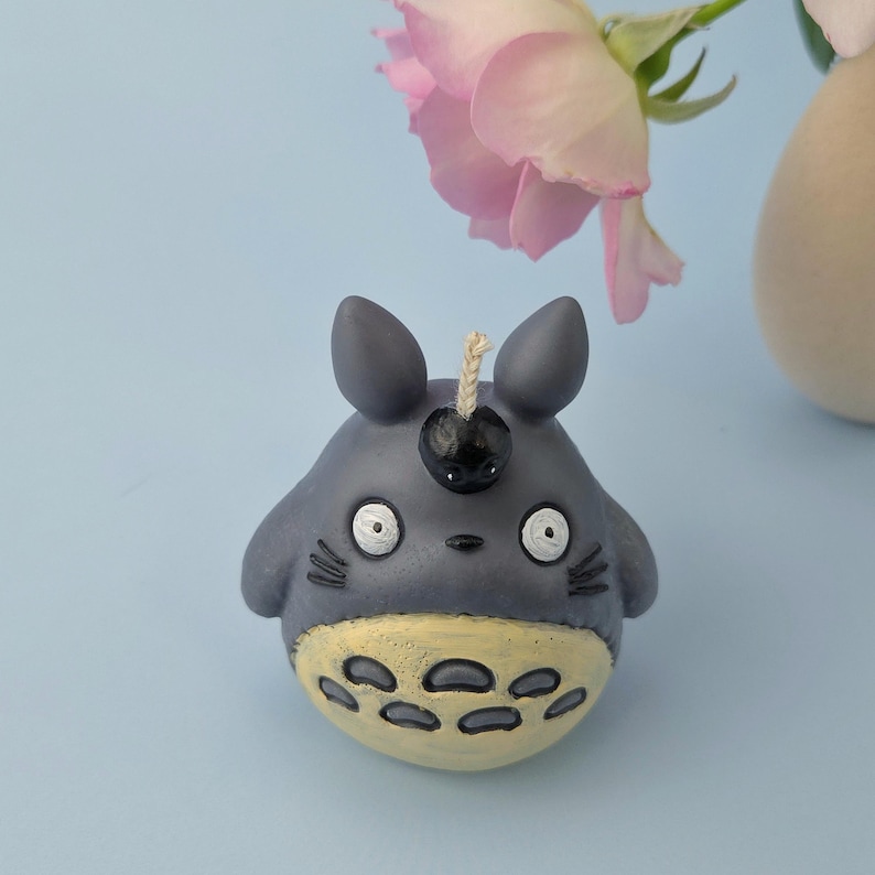 May include: A gray candle shaped like a cartoon character with large eyes, small ears, and a yellow belly. The candle has a black cherry-like detail on its head and a wick. A pink rose and a beige vase are in the background.