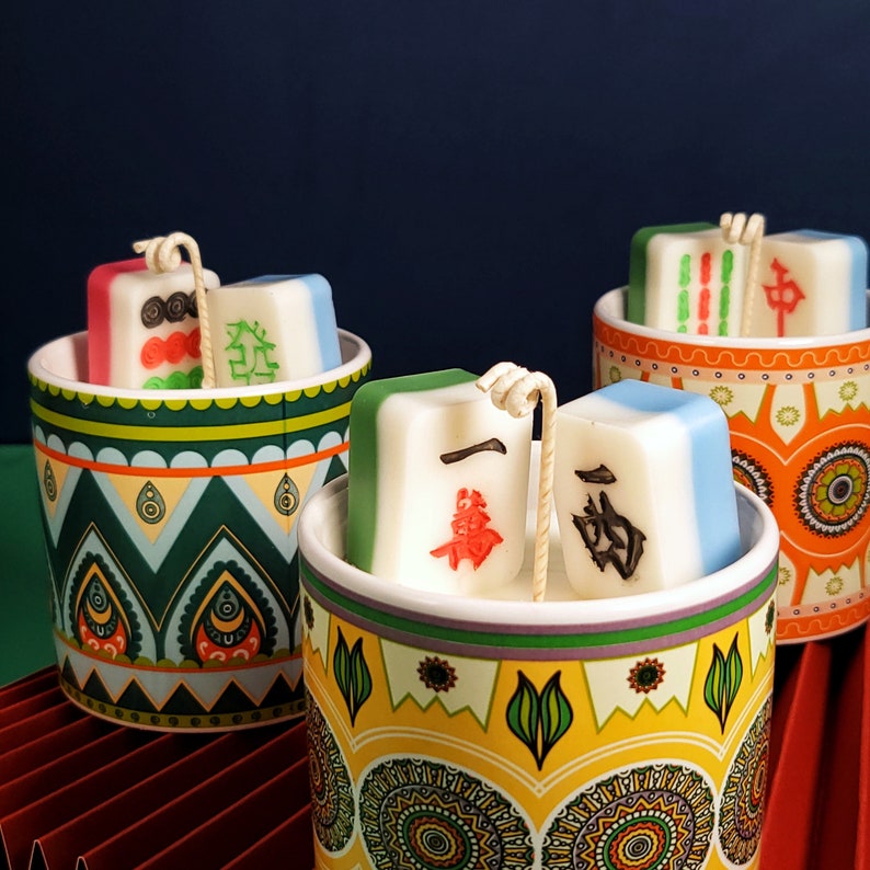 May include: Three ceramic candle holders with colorful patterned designs. Each holder contains a white candle with a green, red, and blue design. The candles are shaped like mahjong tiles.