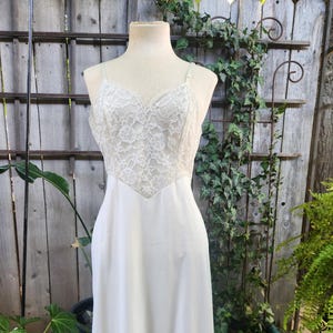 Könnte beinhalten: Ein elfenbeinfarbenes Vintage-Unterkleid auf einer Schaufensterpuppe. Das Oberteil hat eine zarte Spitzenauflage mit V-Ausschnitt und verstellbaren Trägern. Der Rock ist aus glattem Stoff, am Saum mit Spitze besetzt. Das Kleid wird im Freien präsentiert.