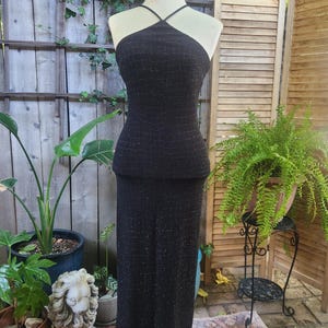 May include: A black, floor-length gown with a halter neckline is displayed on a mannequin. The dress has a textured, shimmering appearance. The mannequin is positioned outdoors, surrounded by plants and a wooden screen.