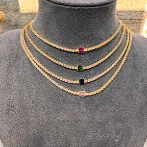 Peut inclure: Quatre colliers dorés avec des pierres claires et des pierres rectangulaires colorées. Les pierres sont roses, bleues, vertes et rouges. Les colliers sont présentés sur un mannequin gris.