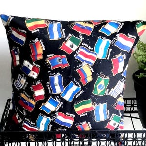 May include: A square throw pillow featuring a black fabric background printed with a repeating pattern of various colorful national flags from Central and South American countries, each accompanied by its respective country name in white text.
