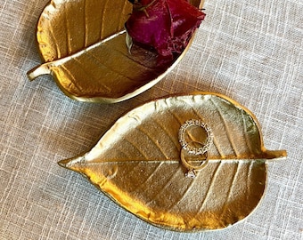 May include: Two gold leaf-shaped trinket dishes. One dish has a dried red rose bud in it. The other dish has a ring with a diamond and a band of small diamonds.