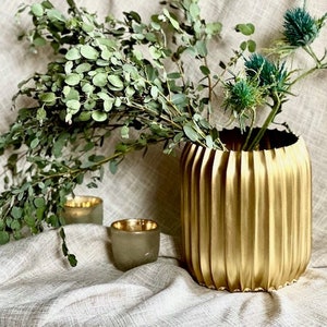 Pode incluir: Um vaso dourado com nervuras com um buquê de flores de eucalipto e cardo verdes. O vaso está sobre uma superfície de tecido castanho claro.