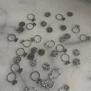 May include: A scattering of silver glitter confetti in the shape of rings. The confetti is on a white marble surface.