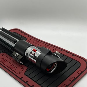May include: A black and silver lightsaber with a red light saber blade. The lightsaber is on a red and black mat.