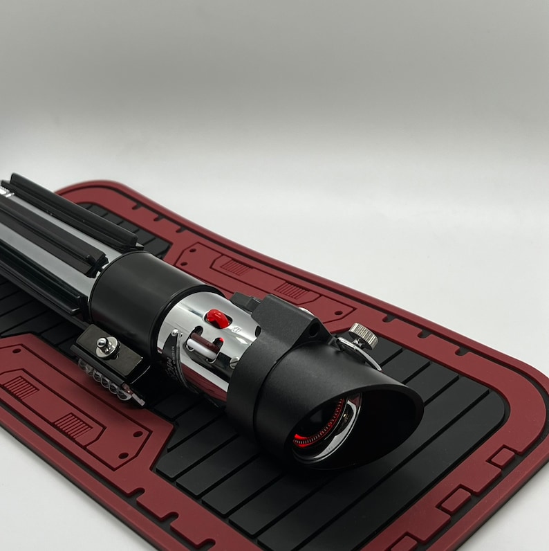May include: A black and silver lightsaber with a red light saber blade. The lightsaber is on a red and black mat.